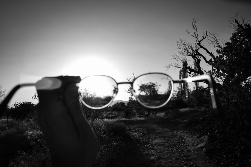Black and white of crop anonymous person holding eyeglasses on walkway in countryside with trees under cloudless sky on sunny summer day